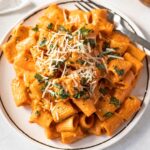 rigatoni pasta with pink sauce on a white plate topped with fresh basil and shredded parmesan cheese.