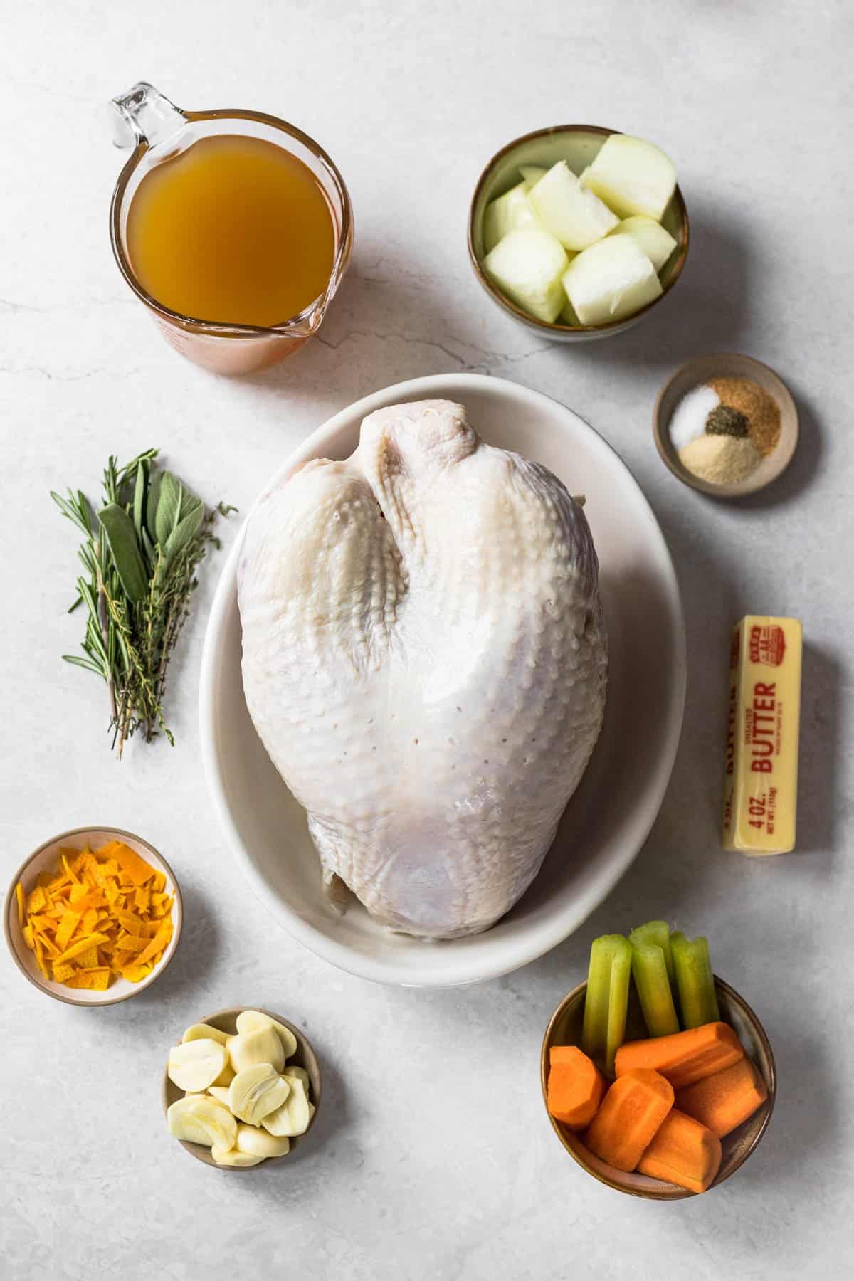 ingredients for bone in turkey breast - turkey broth, whole bone-in breast with skin, quartered onions, seasonings, stick of butter, celery sticks, sliced carrots, garlic cloves, julienned orange peel, rosemary, thyme and sage leaves.