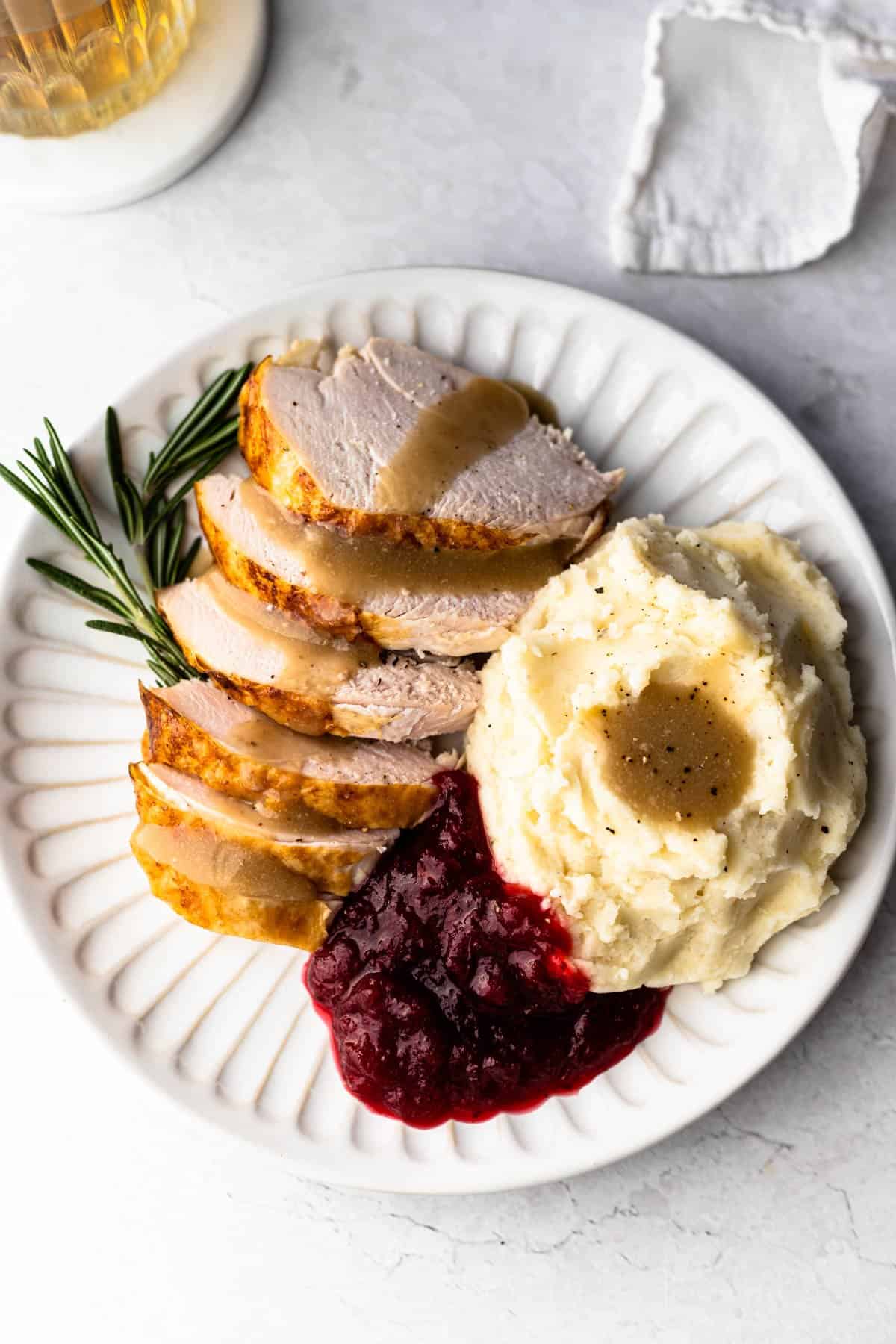 Sliced turkey breast cut into crosswise pieces on a white scalloped plate served with mashed potatoes, gravy and cranberry sauce and a sprig of rosemary.