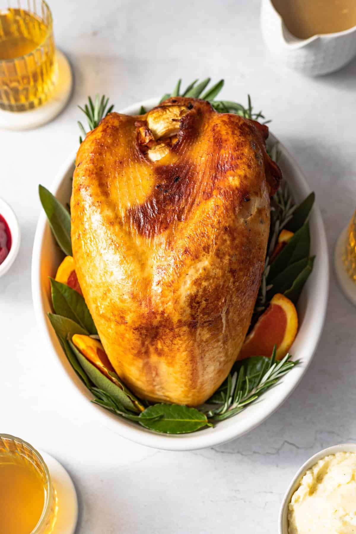 whole bone in turkey breast with golden skin on a white serving dish garnished with orange slices, bay leaves and rosemary.