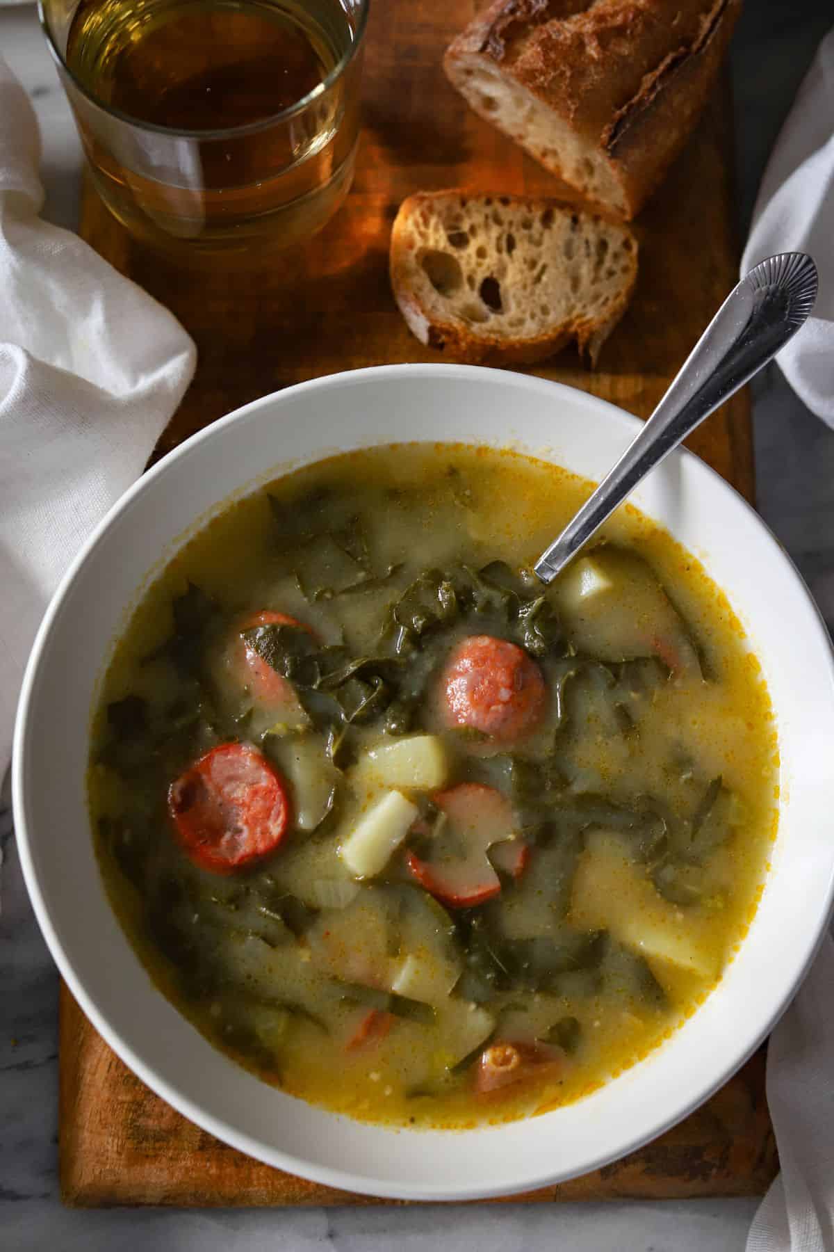 bowl of caldo verde served with a glass of wine and a bread baguette.
