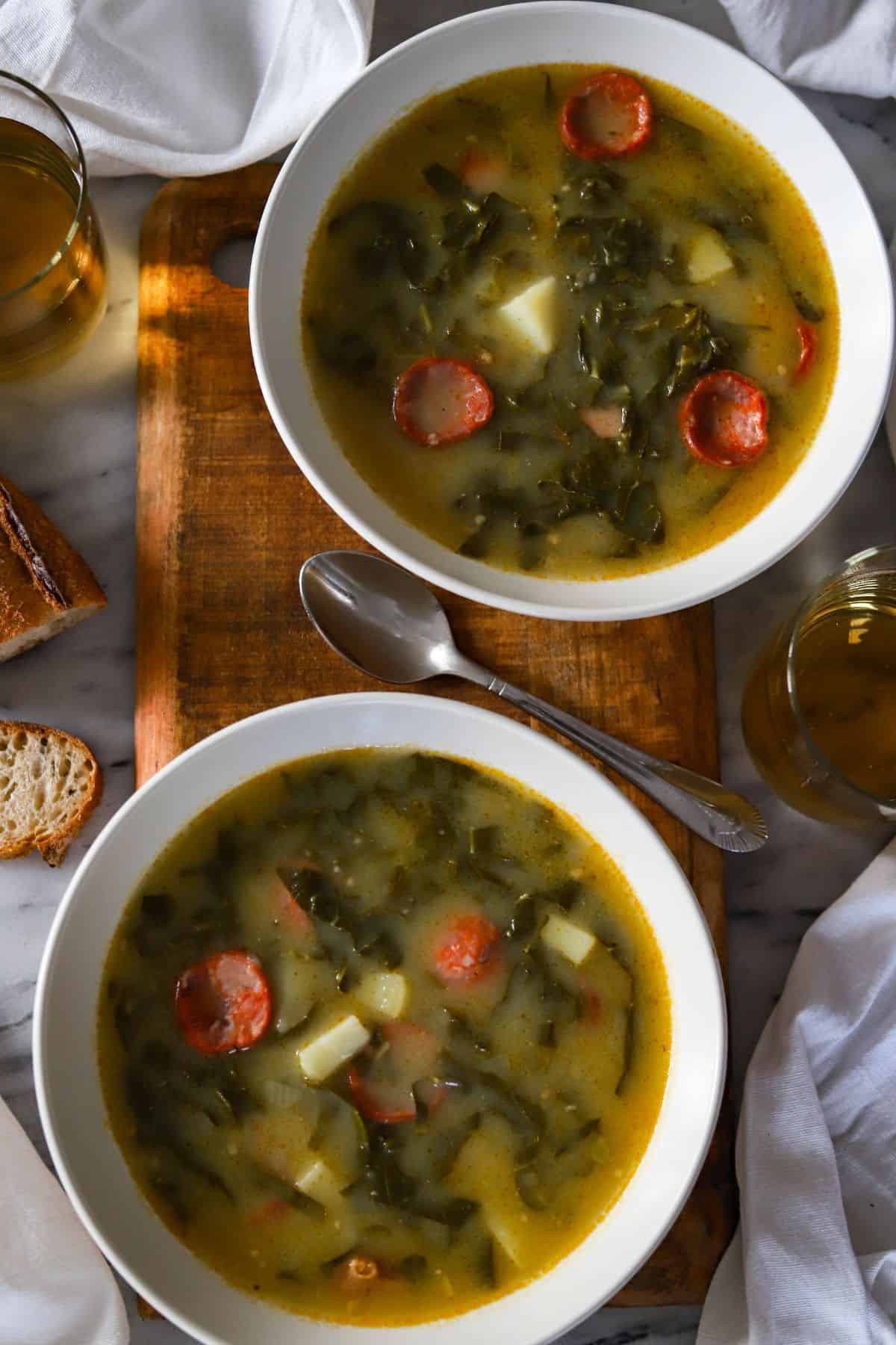 two bowls of caldo verde shown with sliced bread and two glasses of wine.