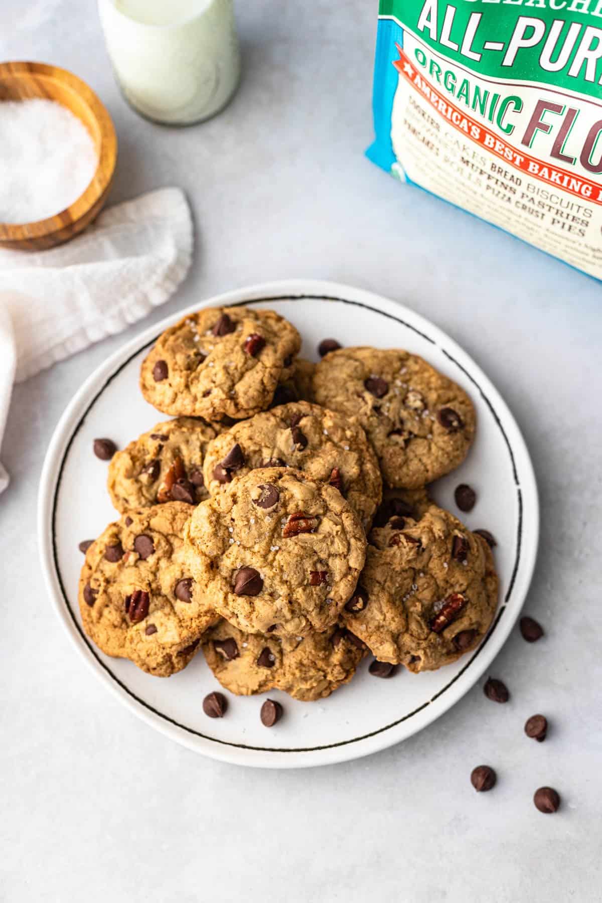 chocolate chip pecan cookies on white plate with bag of flour, bowl sea salt, bottle milk and chocolate chips scattered in background.