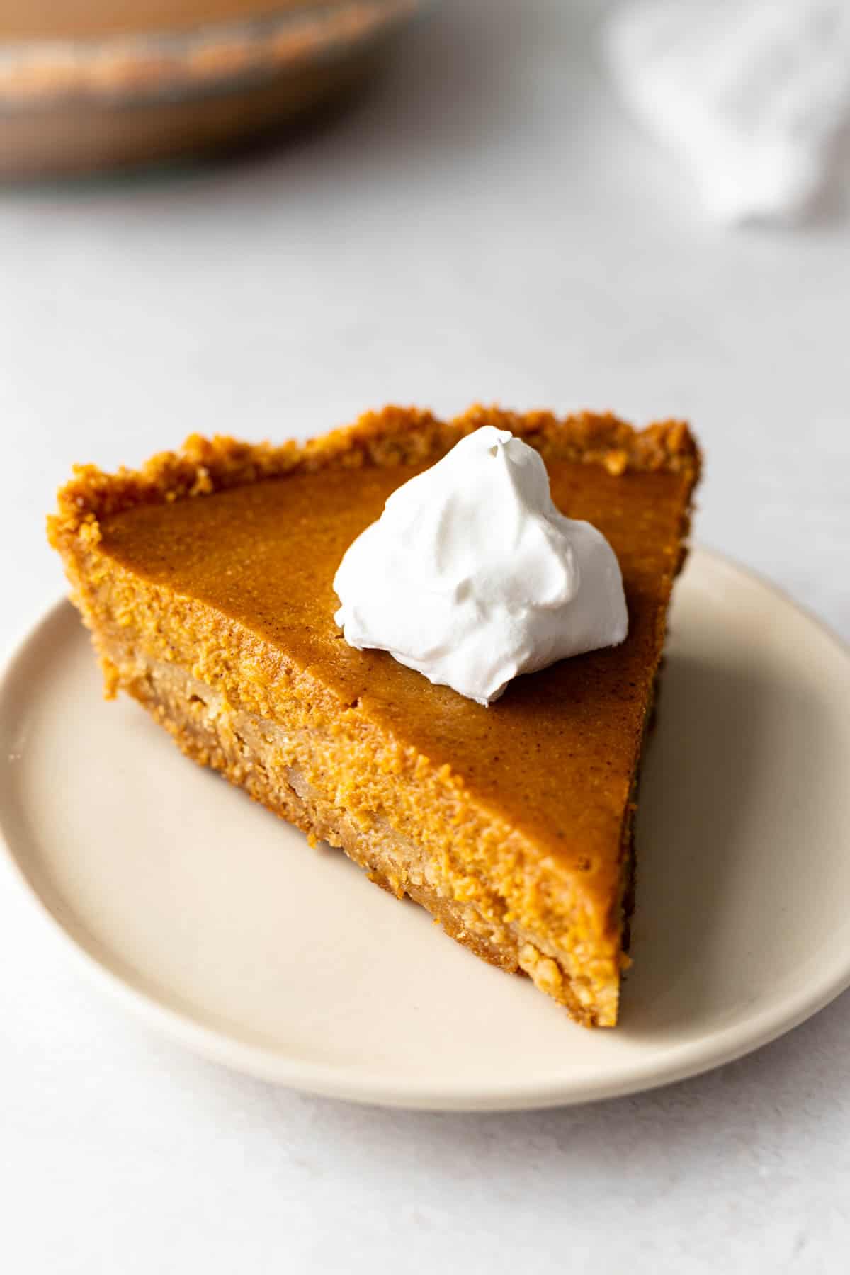 slice of pumpkin pie on a graham cracker crust topped with whipped topping. Full pie is in the background.