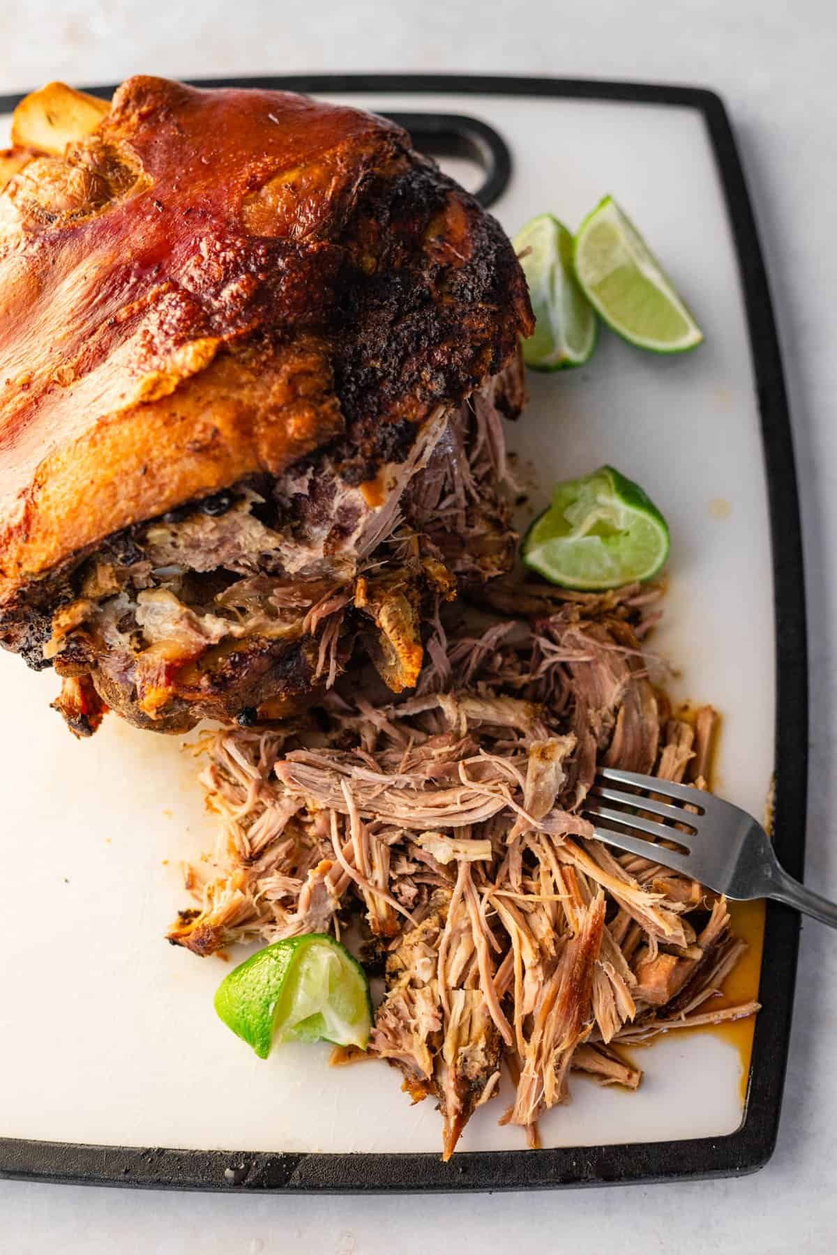 shredded pernil on cutting board shown with skin still in tact and squeezed limes for garnish.