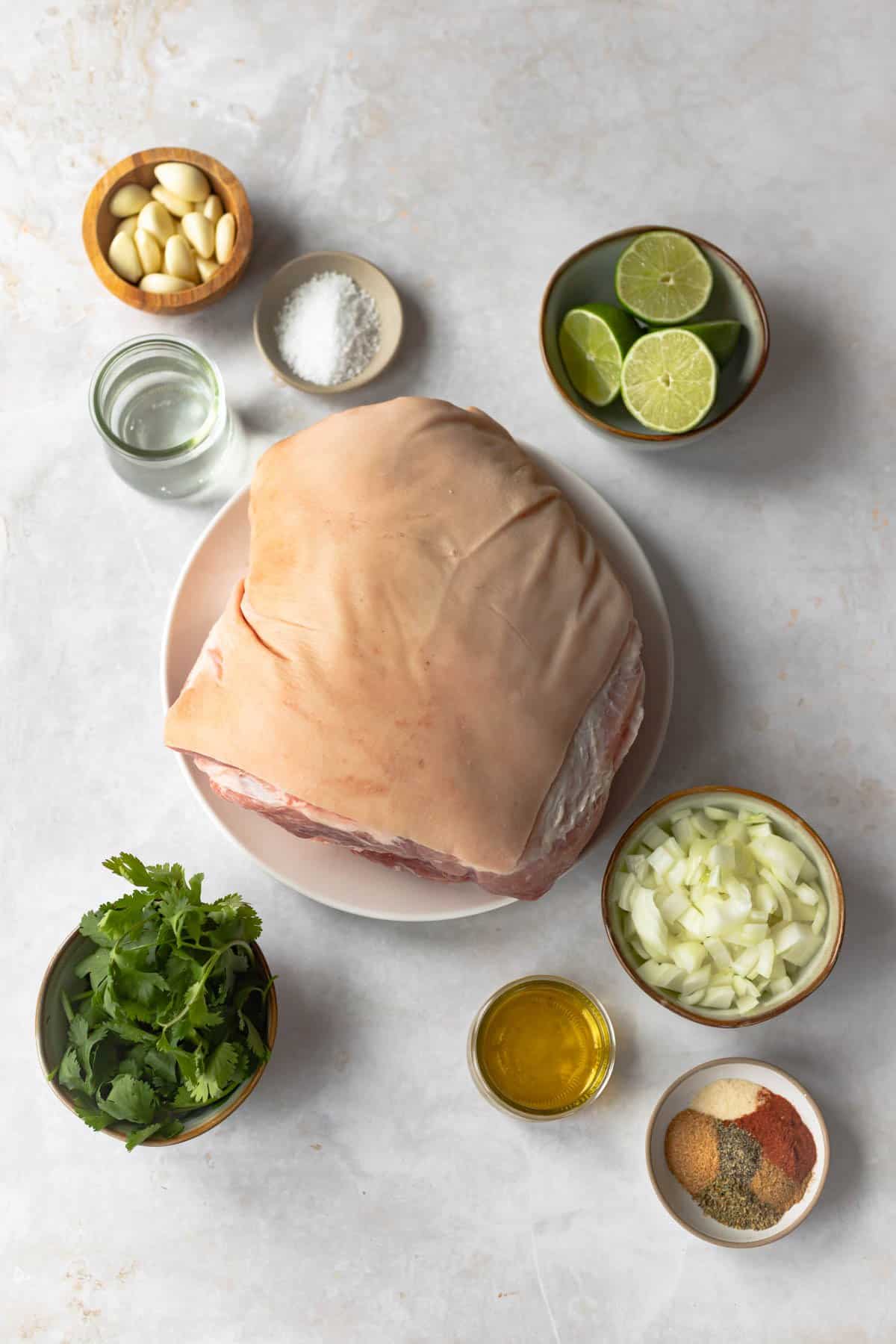 pernil-style roast pork ingredients - garlic, salt, vinegar, fresh limes, picnic roast, cilantro, onions, oil and seasonings.