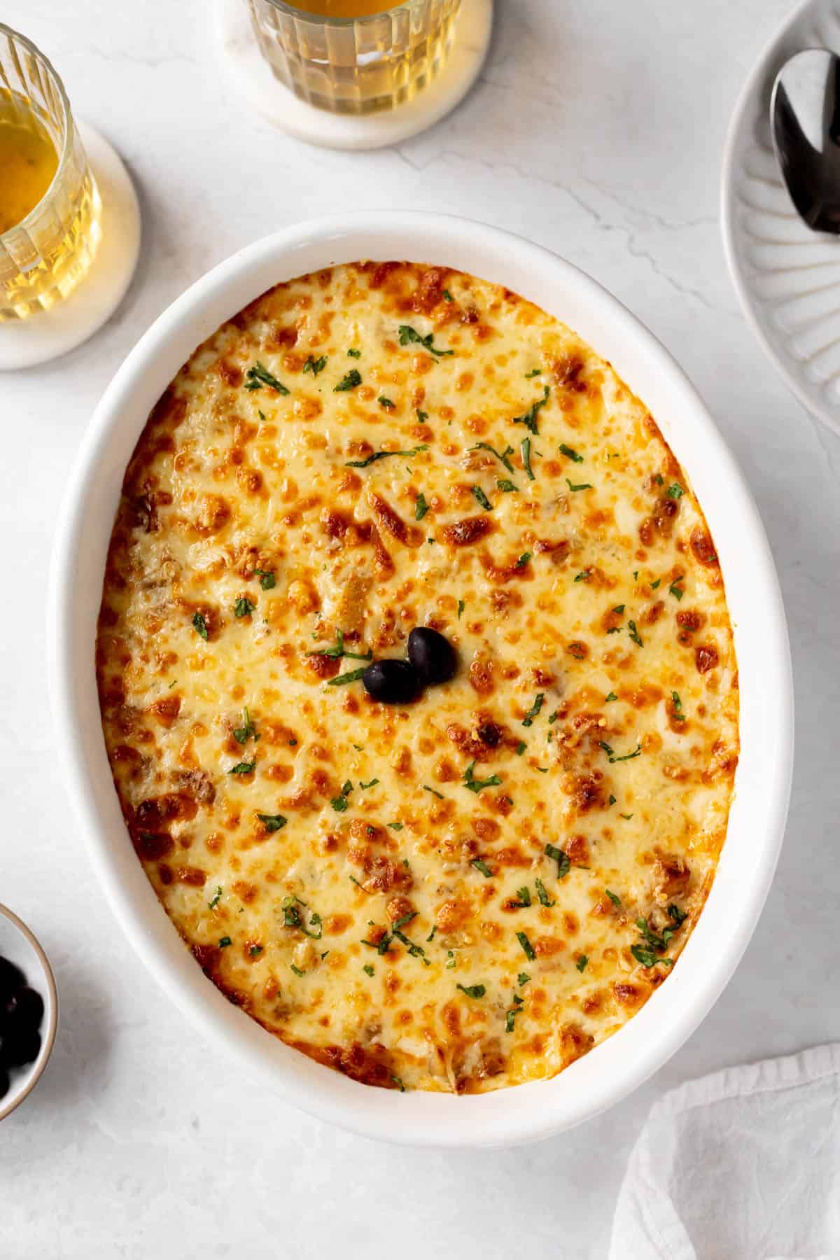 bacalhau com natas in a casserole dish with melted cheese on top and garnished with 2 black olives and chopped parsley.