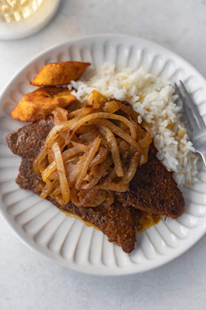 bistec encebollado on a plate with white rice and fried sweet plantains.