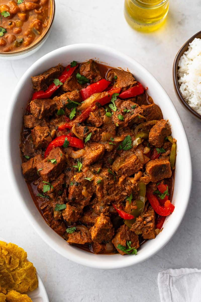 Carne guisada served in a white, oval baking dish, with tostones, white rice and Dominican beans on the side.
