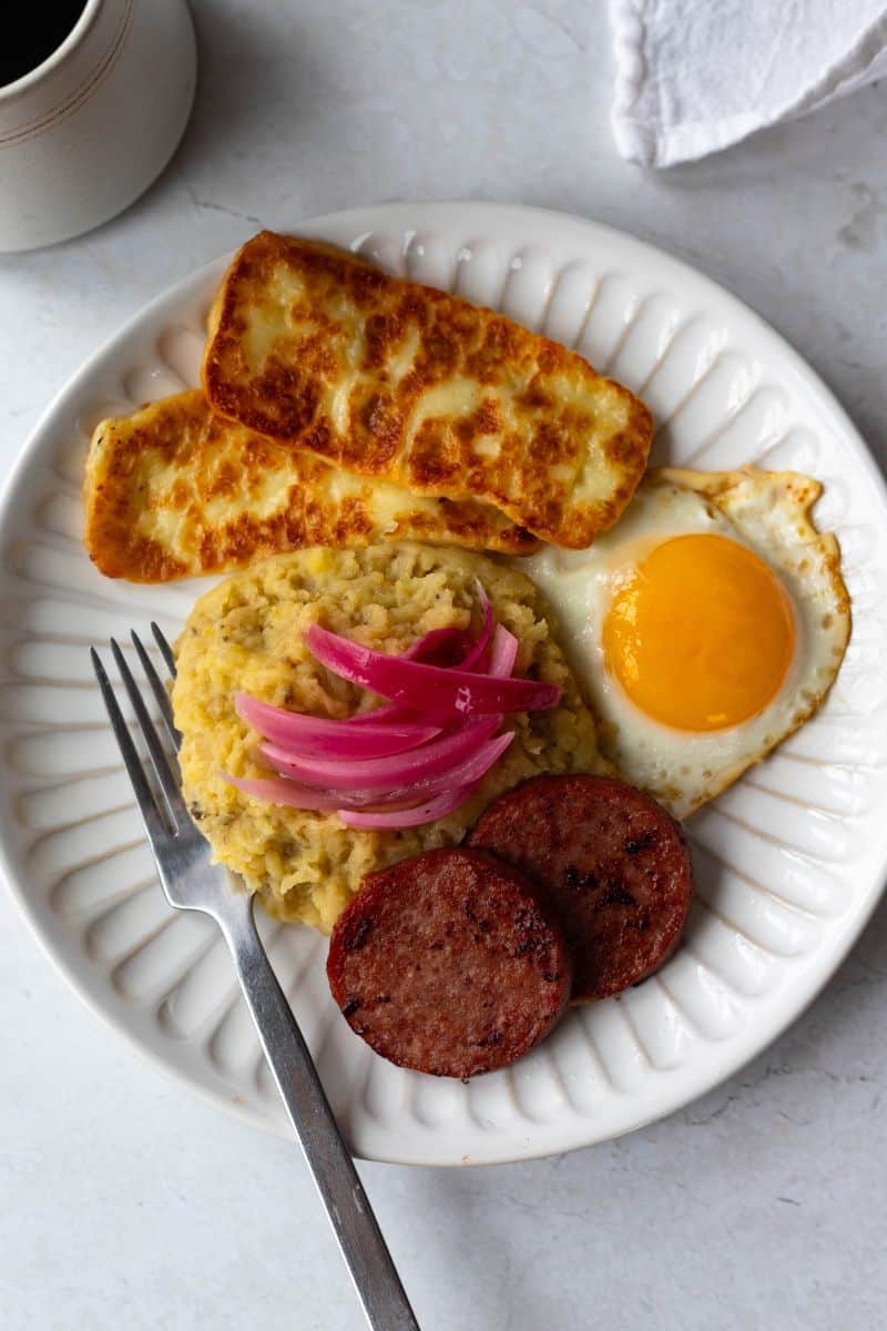 mangu con los tres golpes (salami, fried cheese and eggs) on a white plate, served with a cup of black coffee. 